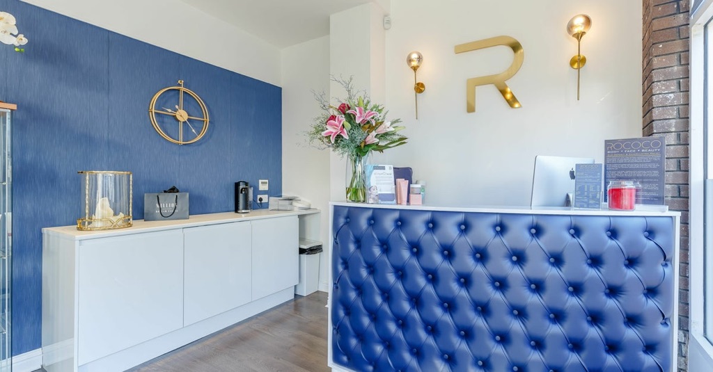 The Rococo clinic reception with a gold R monogram, blue tufted desk and floral arrangement